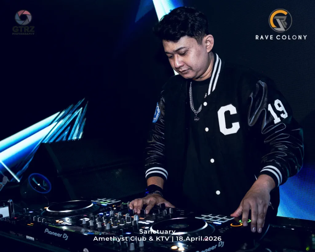 DJ at a nightclub mixing on Pioneer DJ gear, wearing a black varsity jacket, with event text 'Sanctuary Amethyst Club & KTV | 18.April.2026' visible at bottom