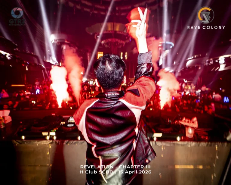 DJ facing away from the camera with raised arms in a neon-lit nightclub, flames and spotlights around, event banner reads 'REVELATION - CHAPTER III' on screen