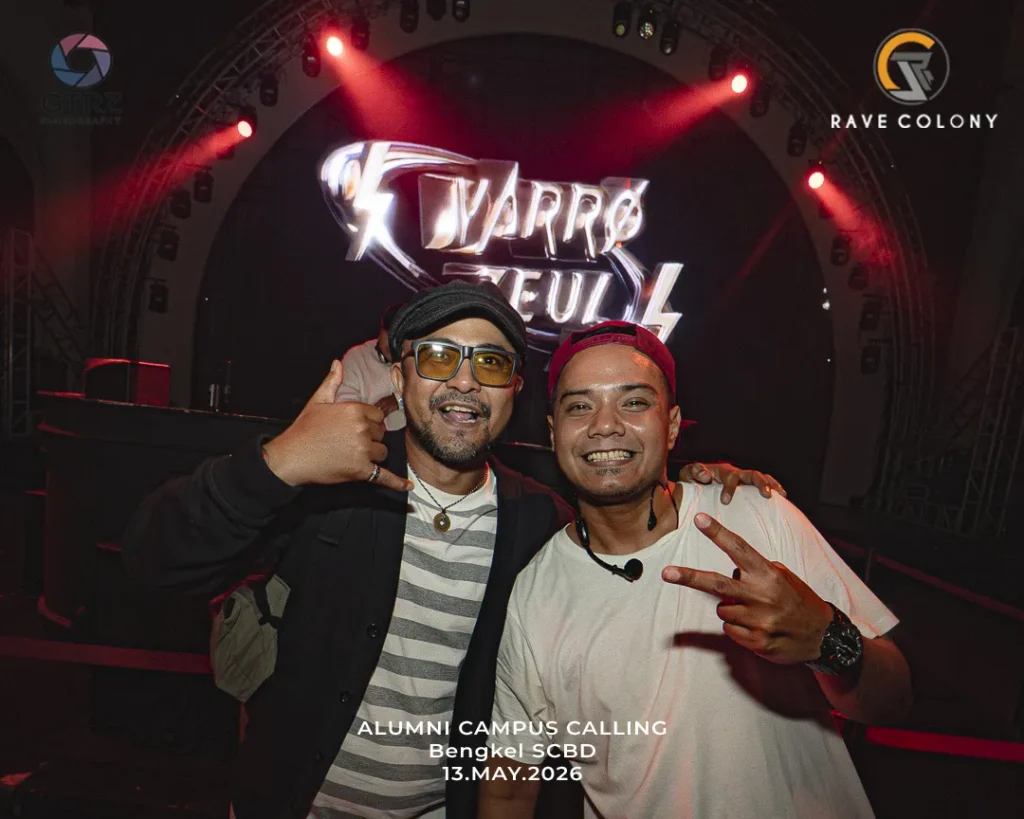 Two men pose together on a dim concert stage, smiling with peace signs, against a bright logo backdrop and event text overlay.