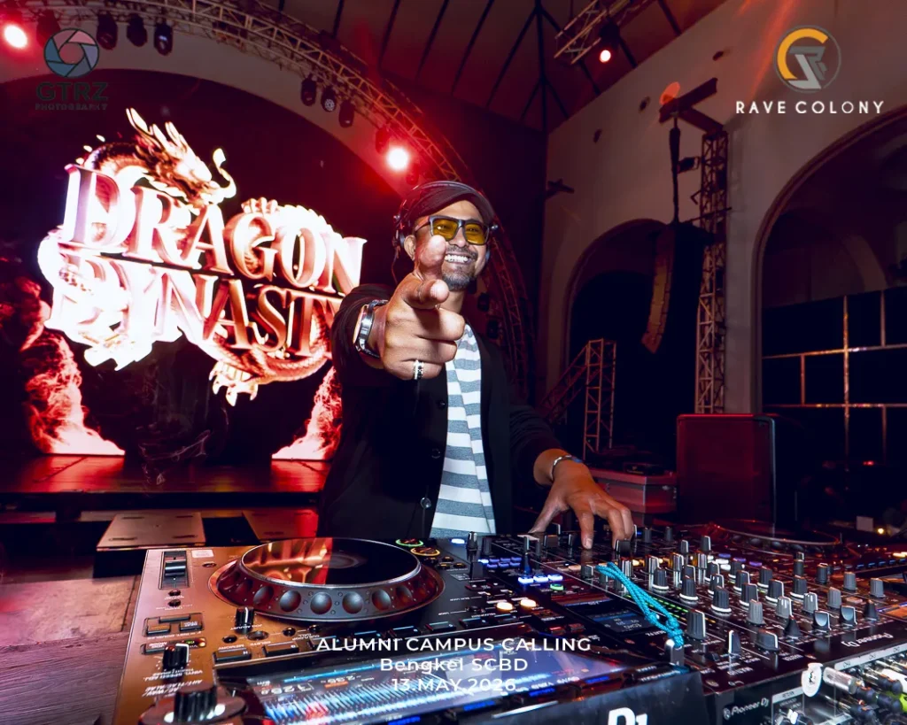 DJ wearing a striped shirt and black jacket points at the camera from behind a professional mixer on a stage with a dragon-themed backdrop and branding nearby.