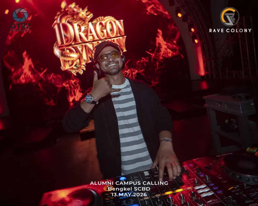DJ at a red-lit club wearing striped shirt and black cardigan, making a thumbs-up/rock gesture while mixing on a DJ console with event graphics in the background.