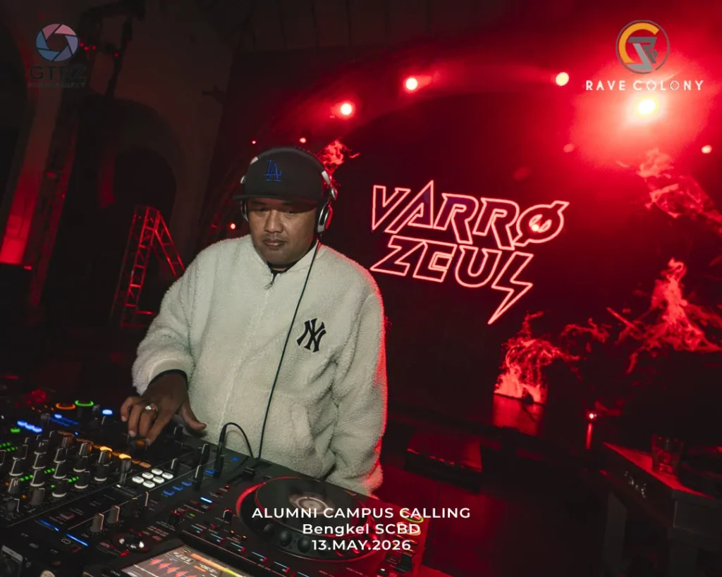 DJ in a white fleece and LA cap mixes on a crowded DJ console under red stage lights, with a neon 'VARRO ZGUL' backdrop and the event text 'ALUMNI CAMPUS CALLING 13 MAY 2026' overlaid in front.