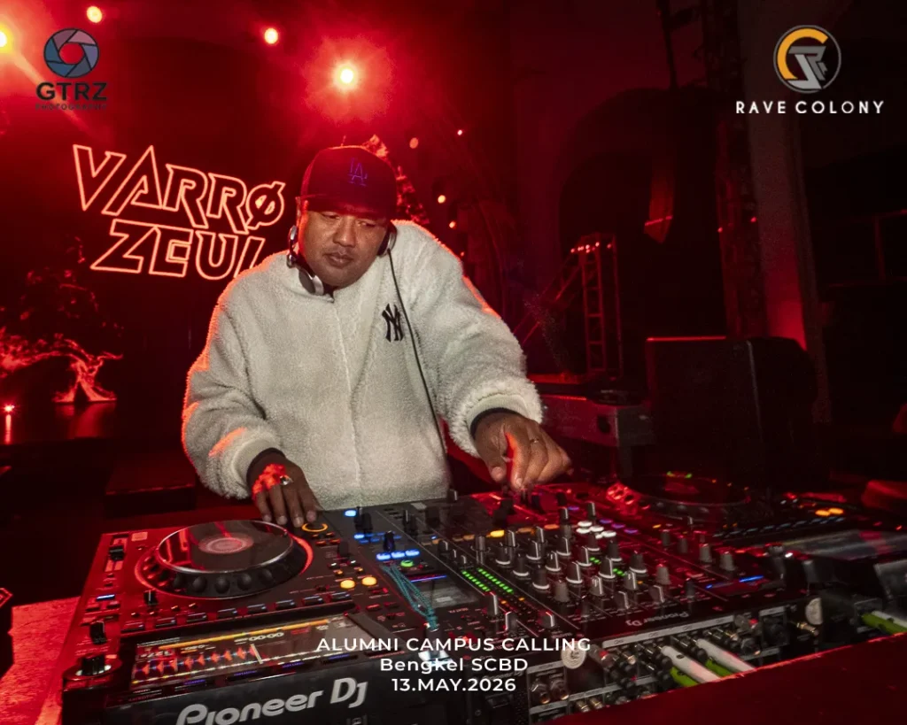 DJ in a white fleece jacket and cap mixes on Pioneer gear under red stage lights at Alumni Campus Calling, Bengkel SCBD, May 13, 2026.