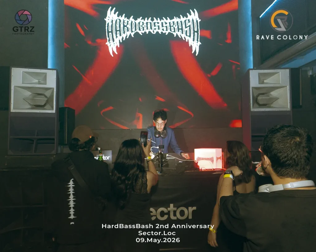 DJ performing on a stage with headphones, large red-lit screen behind, crowd taking photos; banner reads 'HardBassBash 2nd Anniversary Sector.Loc 09.May.2026' with logos on sides