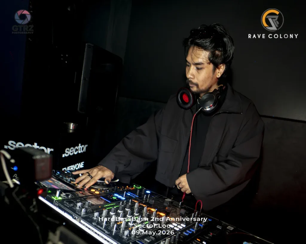 DJ in a black jacket wearing red headphones, adjusting a lit mixer at a dark club during Hard Bass Bash 2nd Anniversary.