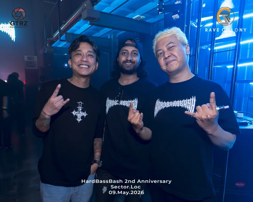 Three men in black shirts pose together at a blue-lit nightclub, smiling and making rock hand signs; event: HardBassBash 2nd Anniversary, 09 May 2026.