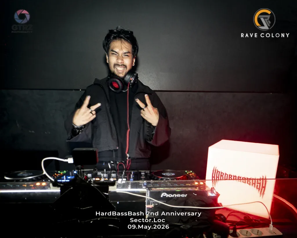DJ posing behind a Pioneer mixer with headphones around neck at HardBassBash 2nd Anniversary, Sector.Loc, May 9, 2026, with logos in the background