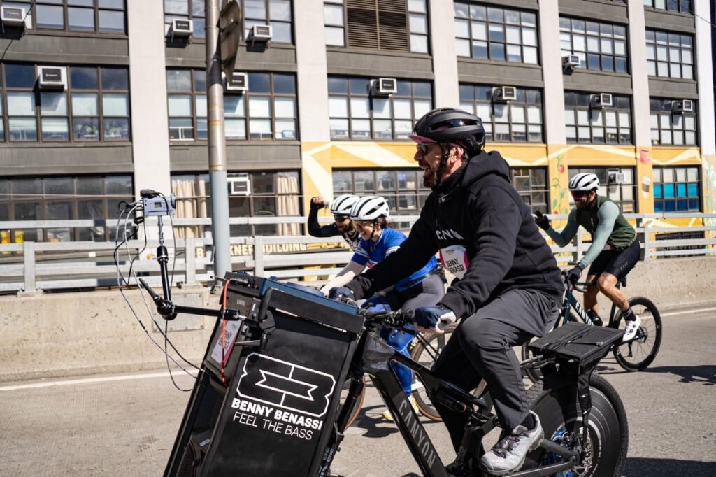 Group of cyclists riding on a city street; the lead bike has Benny Benassi 'Feel the Bass' banner on it.