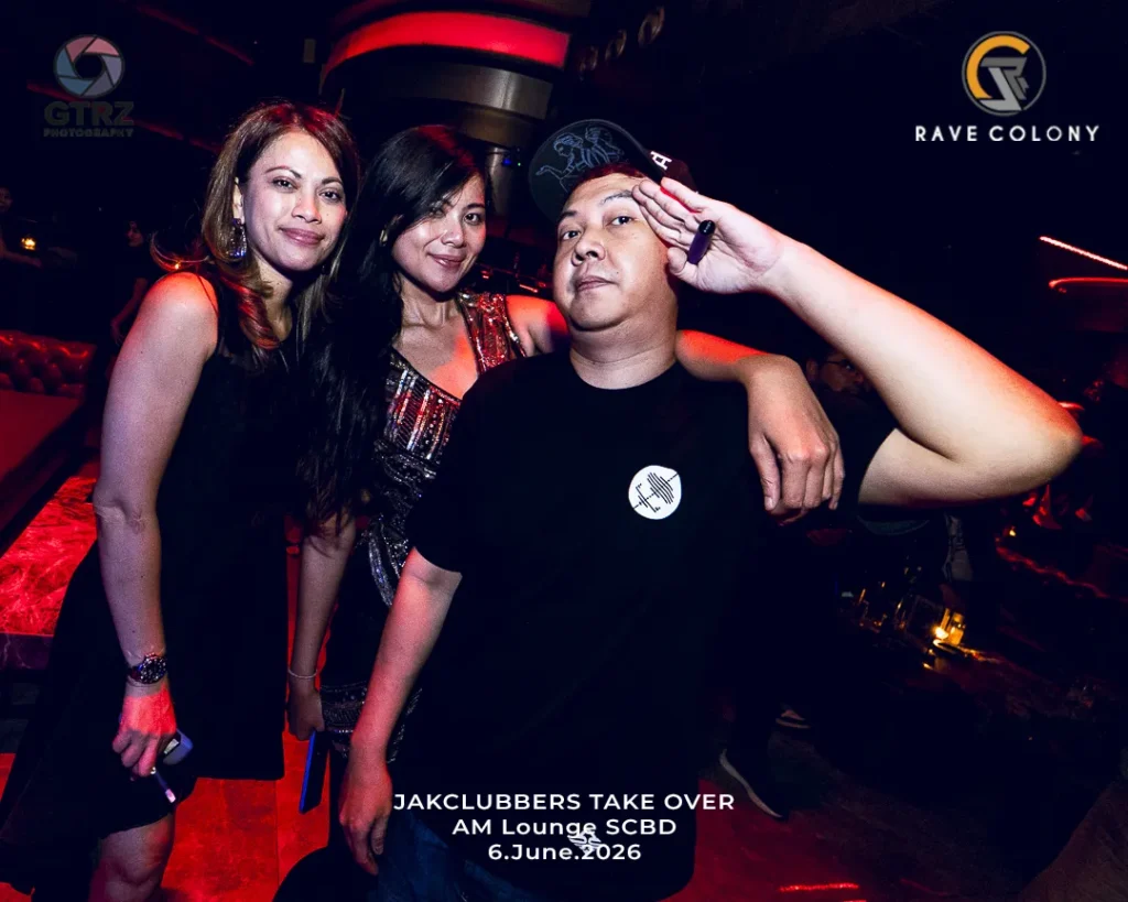 Three people pose in a nightclub: two women on the left and a man in front-right saluting, with colorful club lighting in the background.