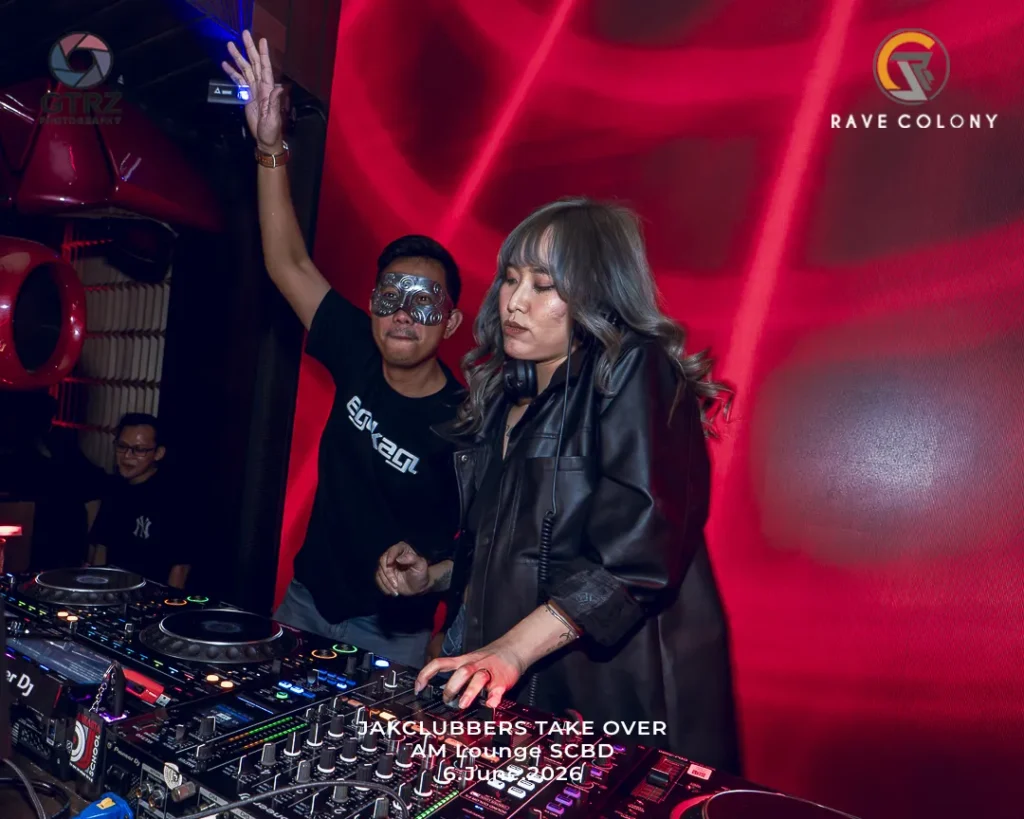 Two club DJs performing at a red-lit venue, woman in front using a mixer while a man wearing a mask raises his hand behind her, with club gear in the foreground.