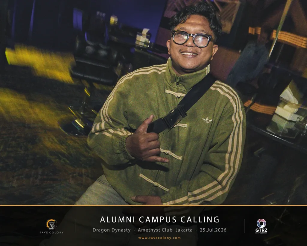 Smiling man with glasses in an olive-green jacket and crossbody bag giving a thumbs-up, in a dim venue with an 'Alumni Campus Calling' banner visible at the bottom.
