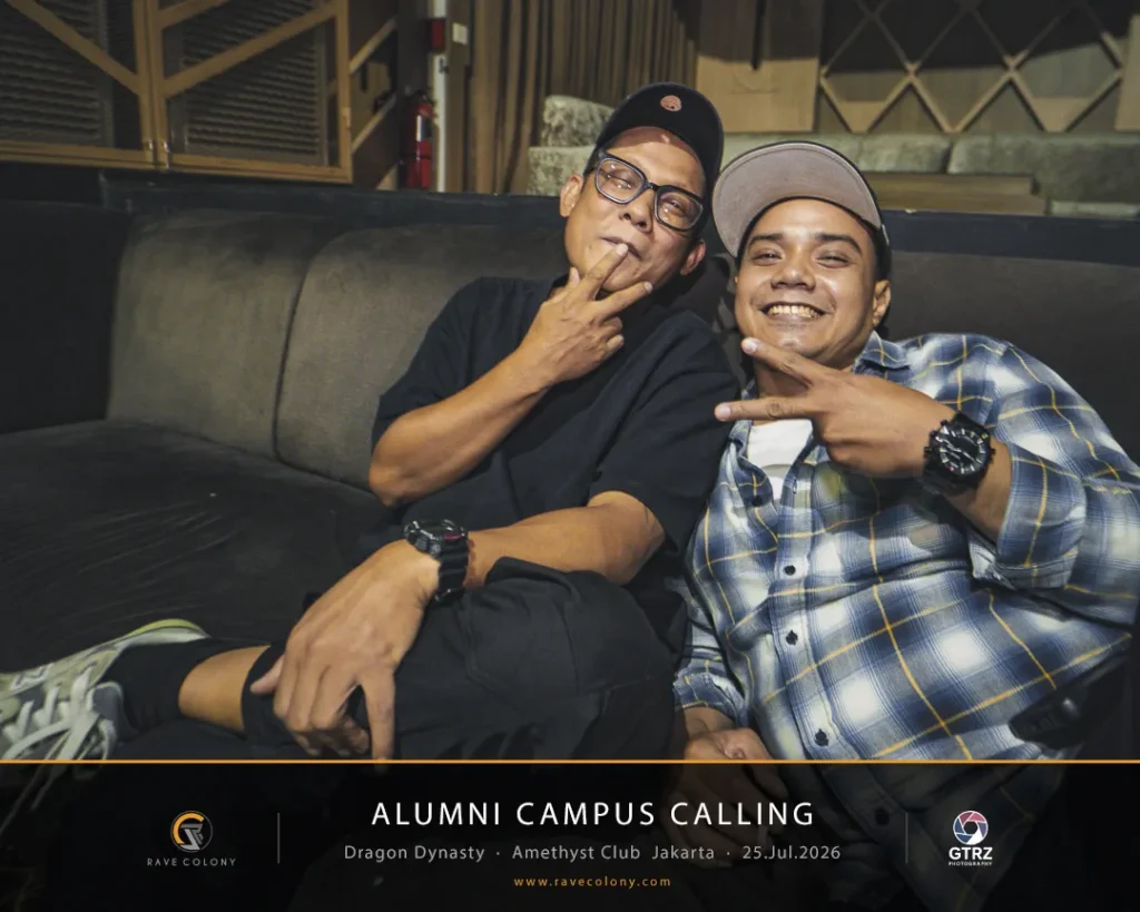 Two men sit on a sofa posing with peace signs at an alumni campus event banner visible below.