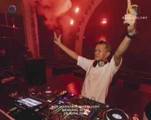 DJ performs on stage at a red lit club raising hands with headphones around neck and fed up with a mixer setup in front