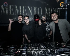 Five friends pose and smile behind a DJ booth in a dim club one wearing a black mask with a red visor at center