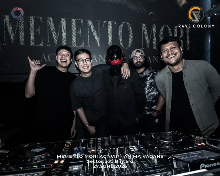 Five friends pose and smile behind a DJ booth in a dim club, one wearing a black mask with a red visor at center.