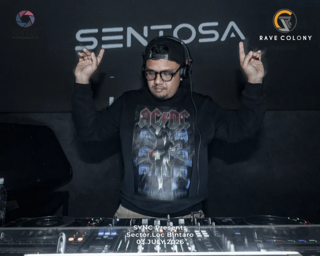 DJ wearing headphones, raising both hands on a nightclub booth with a SENTOSA backdrop and logos on either side, wearing an AC/DC shirt.