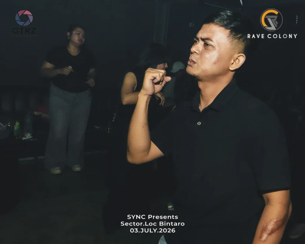 Man in a black polo at a dim nightclub, raising a fist, with event text and logos in the background (SYNC Presents, Sector.Loc Bintaro, 03 July 2026).