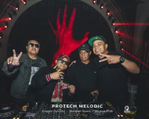 Four men posing at a DJ booth during a night event, with red stage lighting and a large hand graphic in the background.