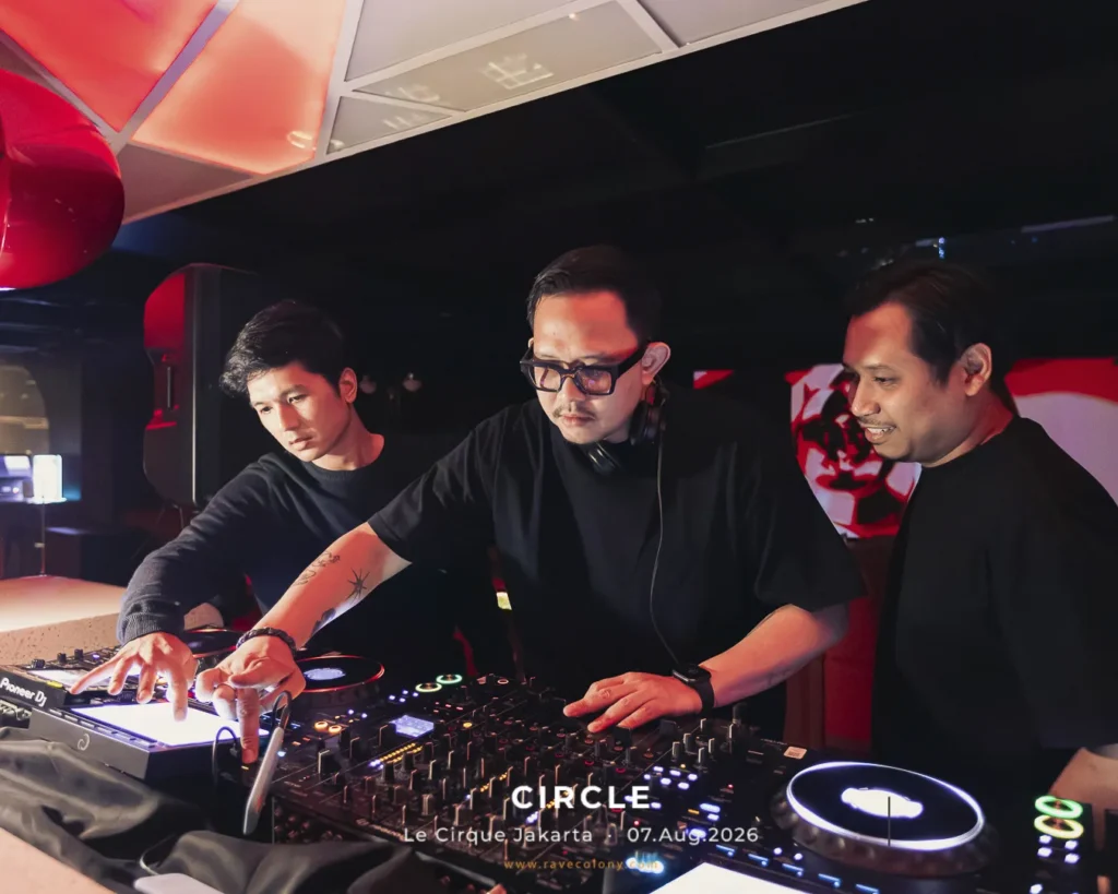 Three DJs in black shirts perform at a club, one wearing glasses and headphones, operating a mixer and turntables under red lighting.