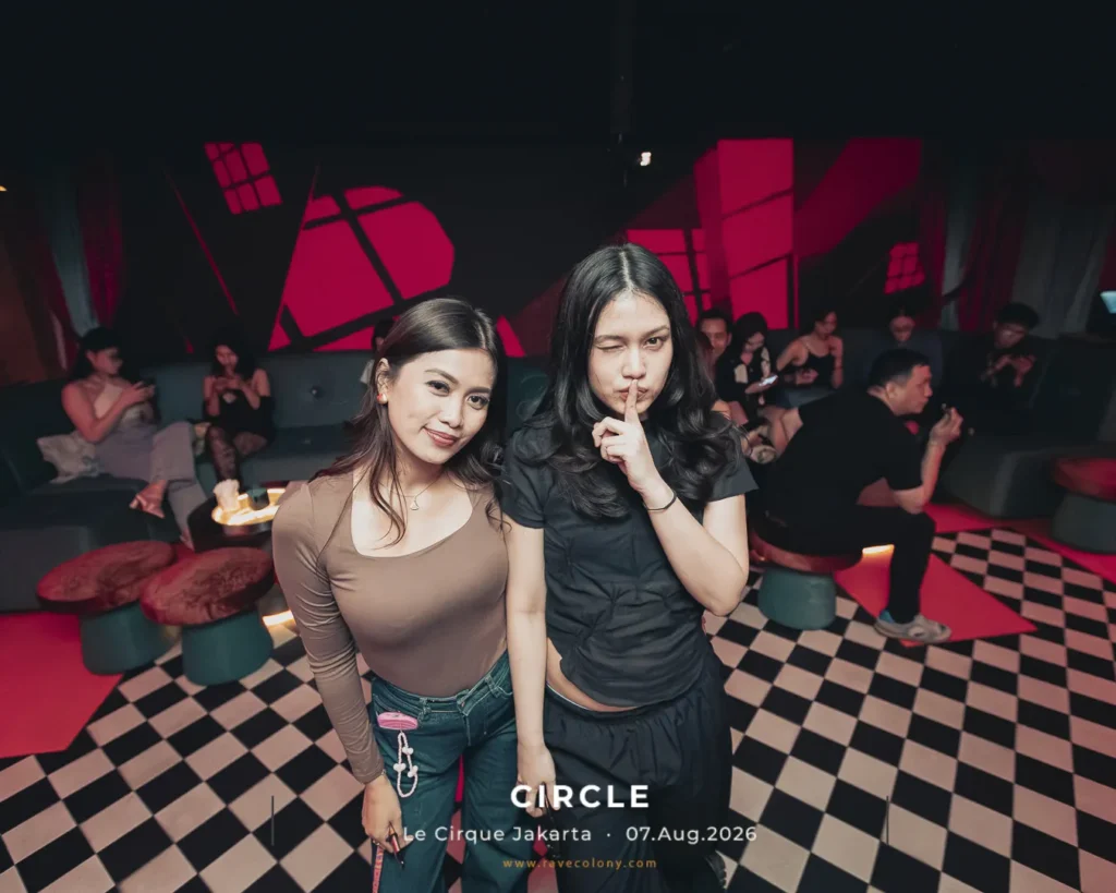 Two young women pose for a photo in a nightclub, one winking and the other making a shushing gesture, with a checkered floor and pink geometric wall art behind them.