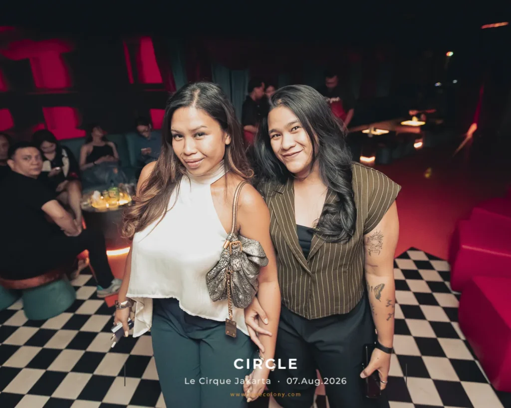 Two women posing and smiling for the camera in a dim club with a black-and-white checkered floor and pink seating in the background.