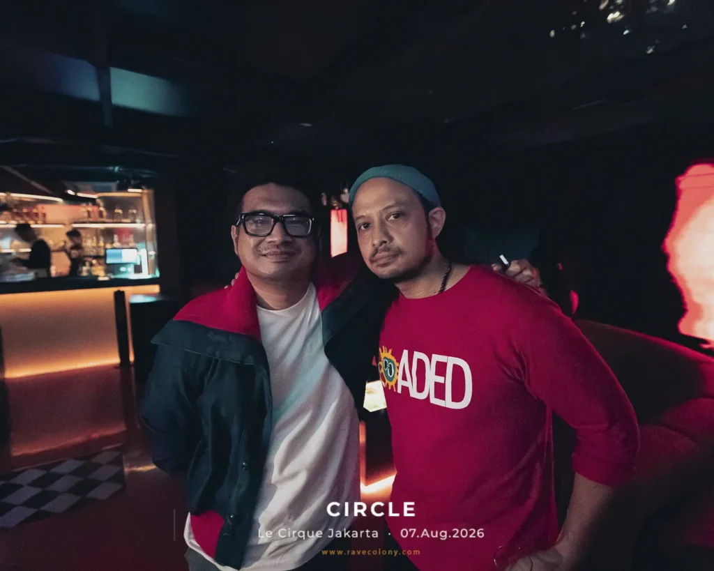 Two men posing together in a dim club, one wearing a red and black jacket and the other a red shirt; warm bar lighting in the background.
