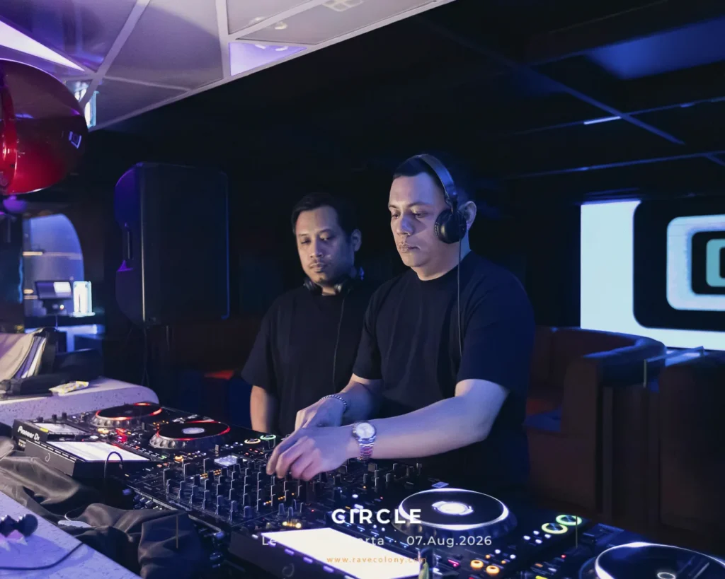 Two male DJs in black shirts perform at a nightclub, adjusting the mixer and turntables under purple lighting.
