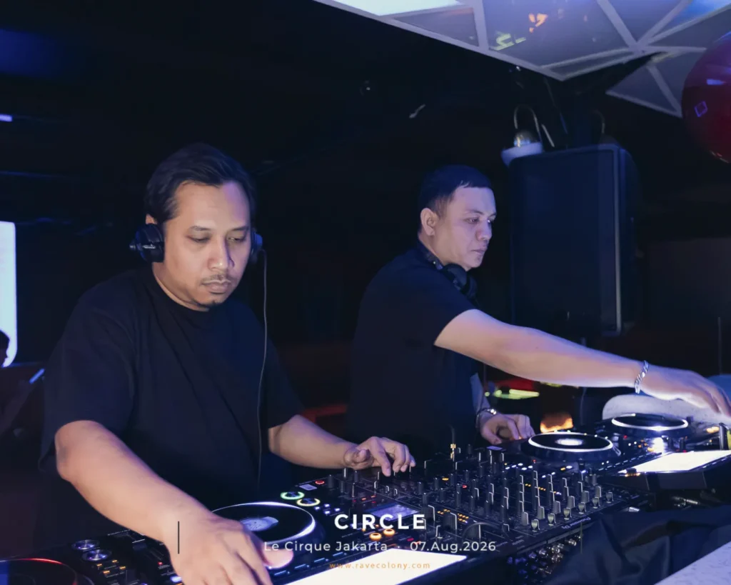 Two DJs perform at a nightclub, wearing black shirts and headphones, mixing tracks on a large DJ console.