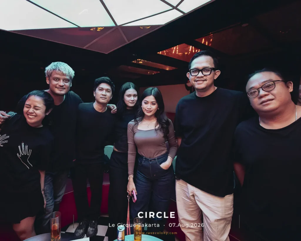 Group of friends posing for a photo in a dark club, smiling and dressed in black, with a checkered floor and drinks on the table in front.