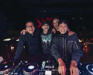 Four men pose and smile behind a DJ booth with turntables in a dim nightclub setting.