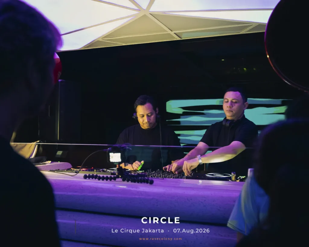 Two DJs mix tracks at a nightclub booth with colorful lighting and audience silhouettes in the foreground.