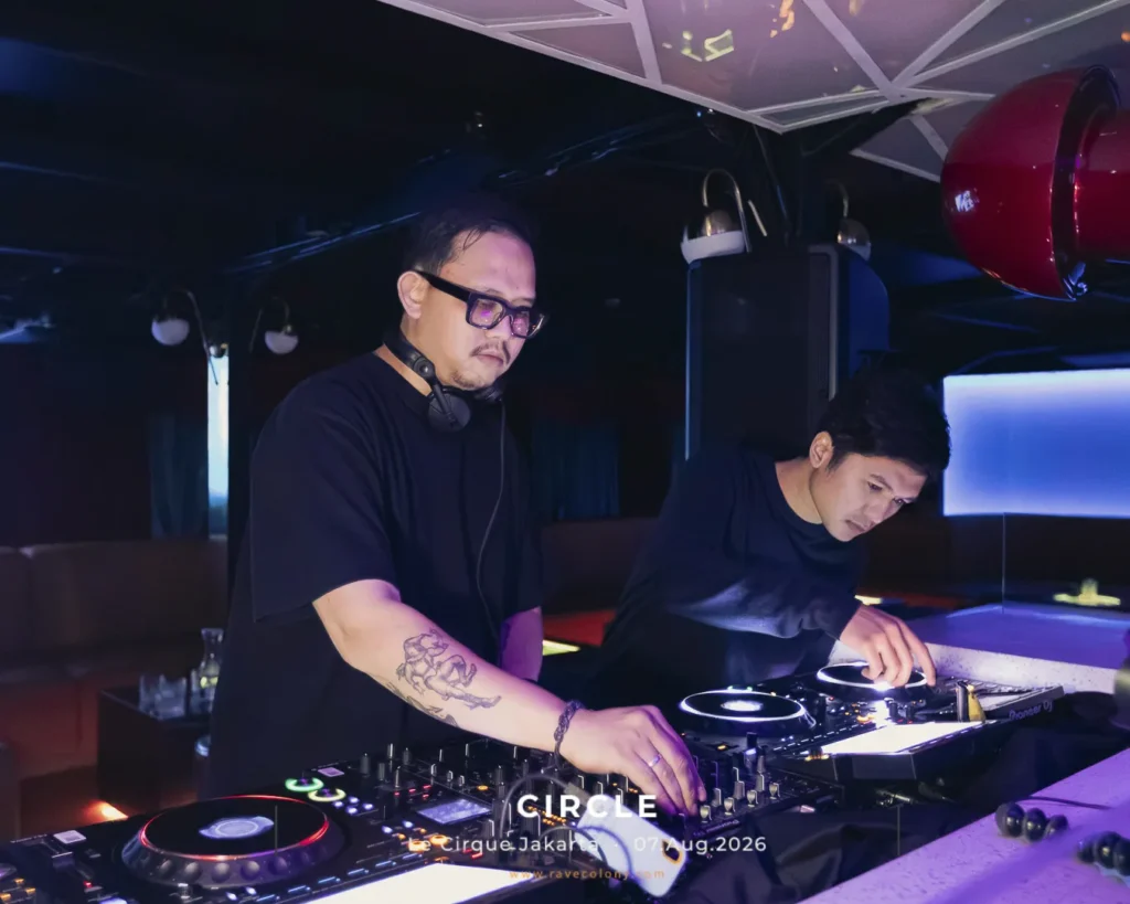 Two male DJs perform at a nightclub, one wearing headphones and adjusting a mixer under blue-purple lighting.