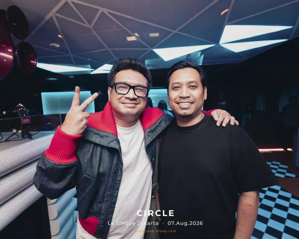 Two men pose together in a nightclub, smiling at the camera, one making a peace sign, with neon lights in the background.